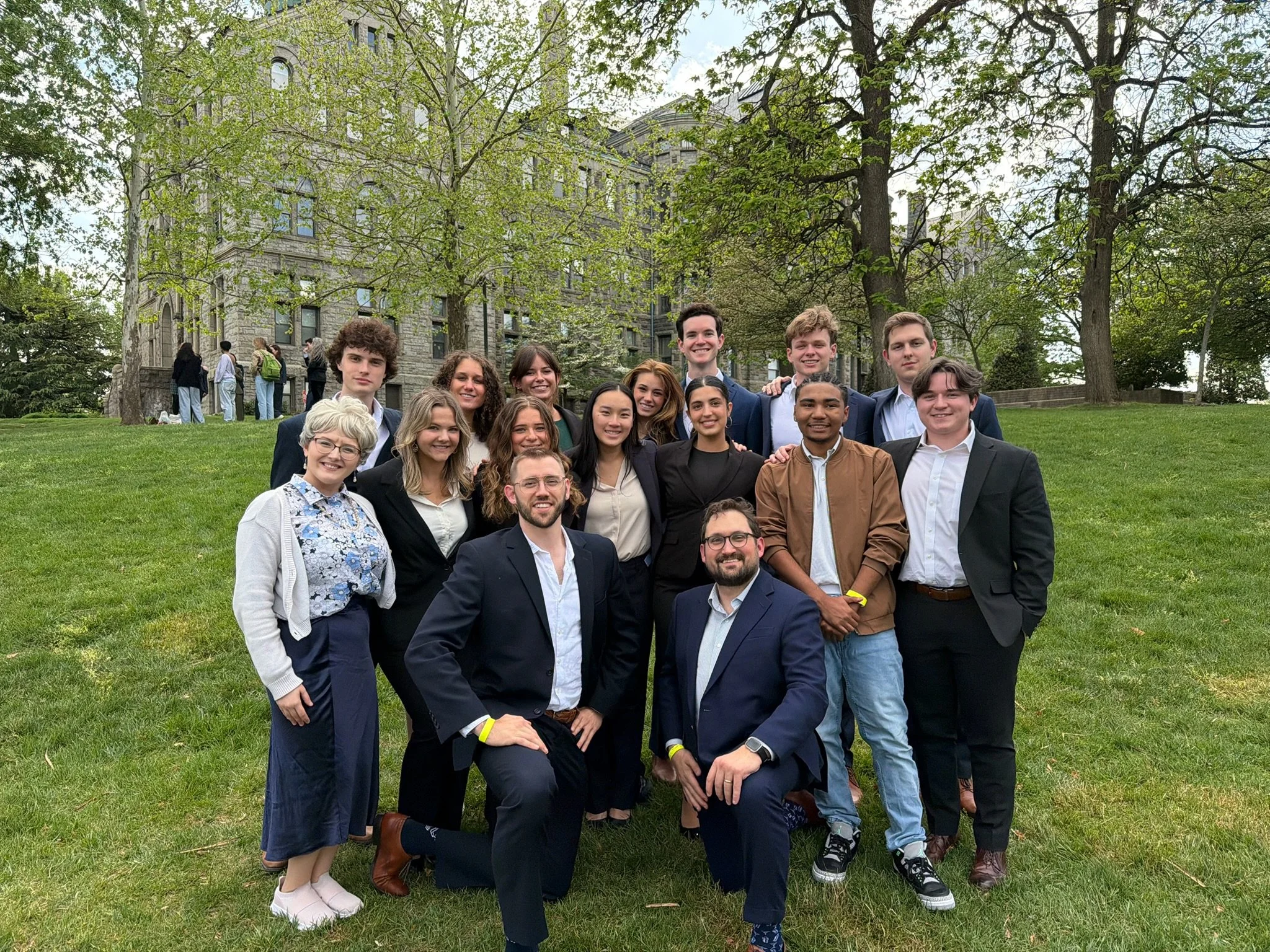 A group of young adults, dressed in formal and casual attire, posing outdoors on a grassy area with trees and a stone building in the background.