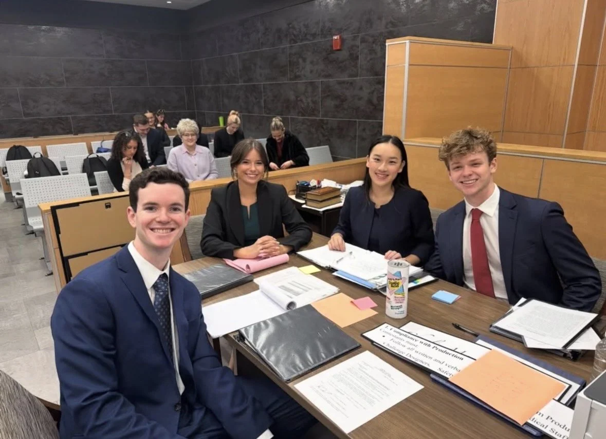 Four young professionals sitting at a table in a courtroom or conference room, smiling and looking at the camera. Behind them, other people are seated and working.