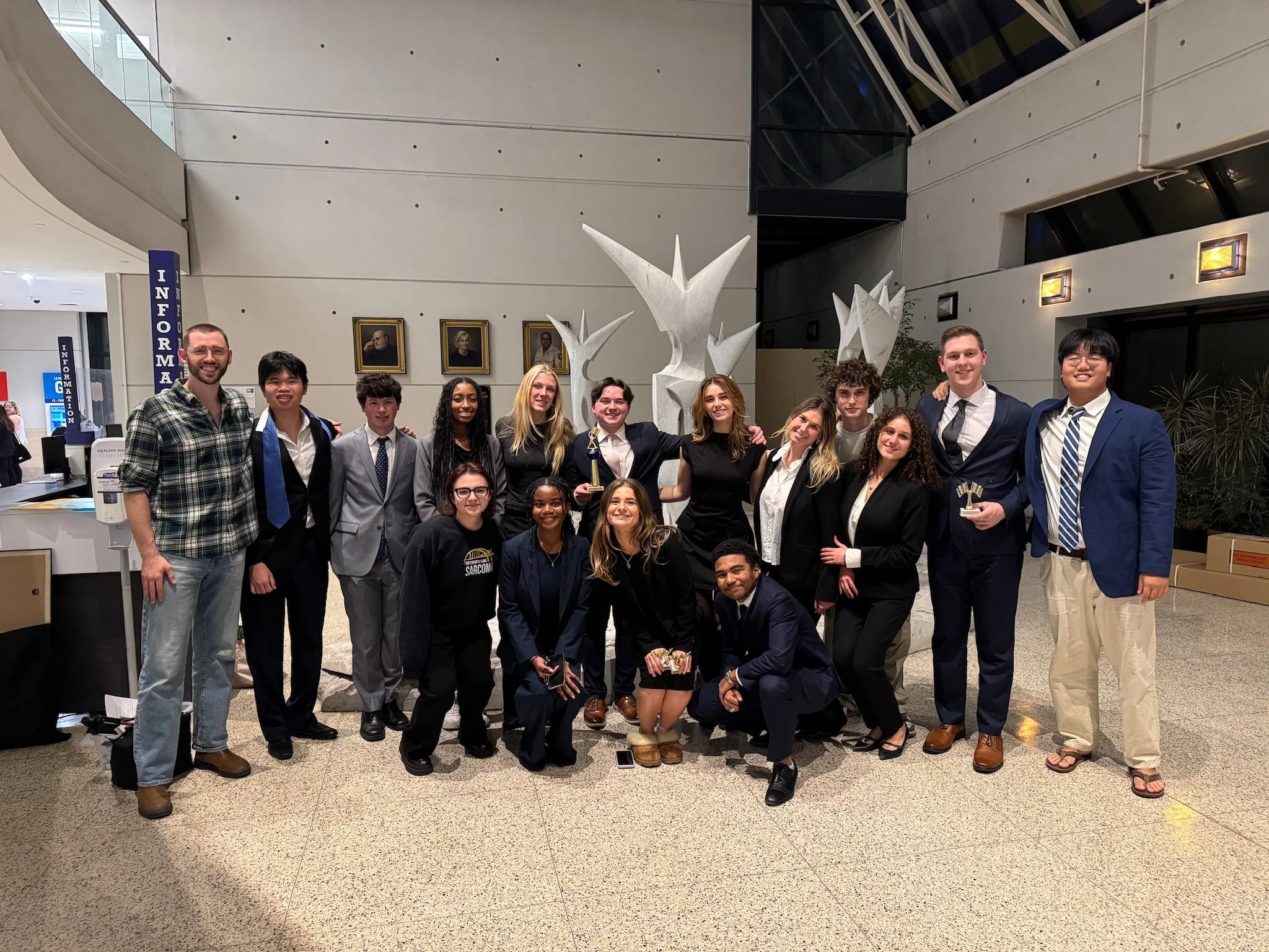 Group of people, mostly young adults, standing together in an indoor space, smiling, dressed in semi-formal attire. Some are holding awards or trophies. The background includes framed portraits, a large abstract sculpture, and area signs marked 'INFORMATION'.