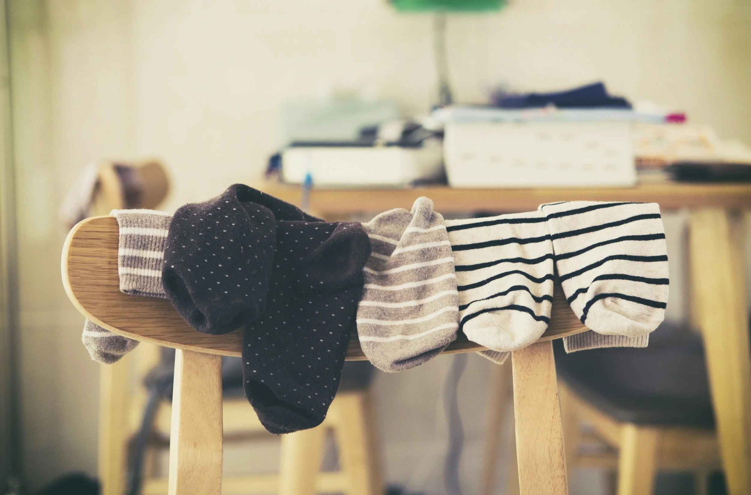 A wooden chair with various socks draped over its backrest, including dark, striped, and patterned socks. In the background, there are stacks of paper and boxes on a table, slightly out of focus.