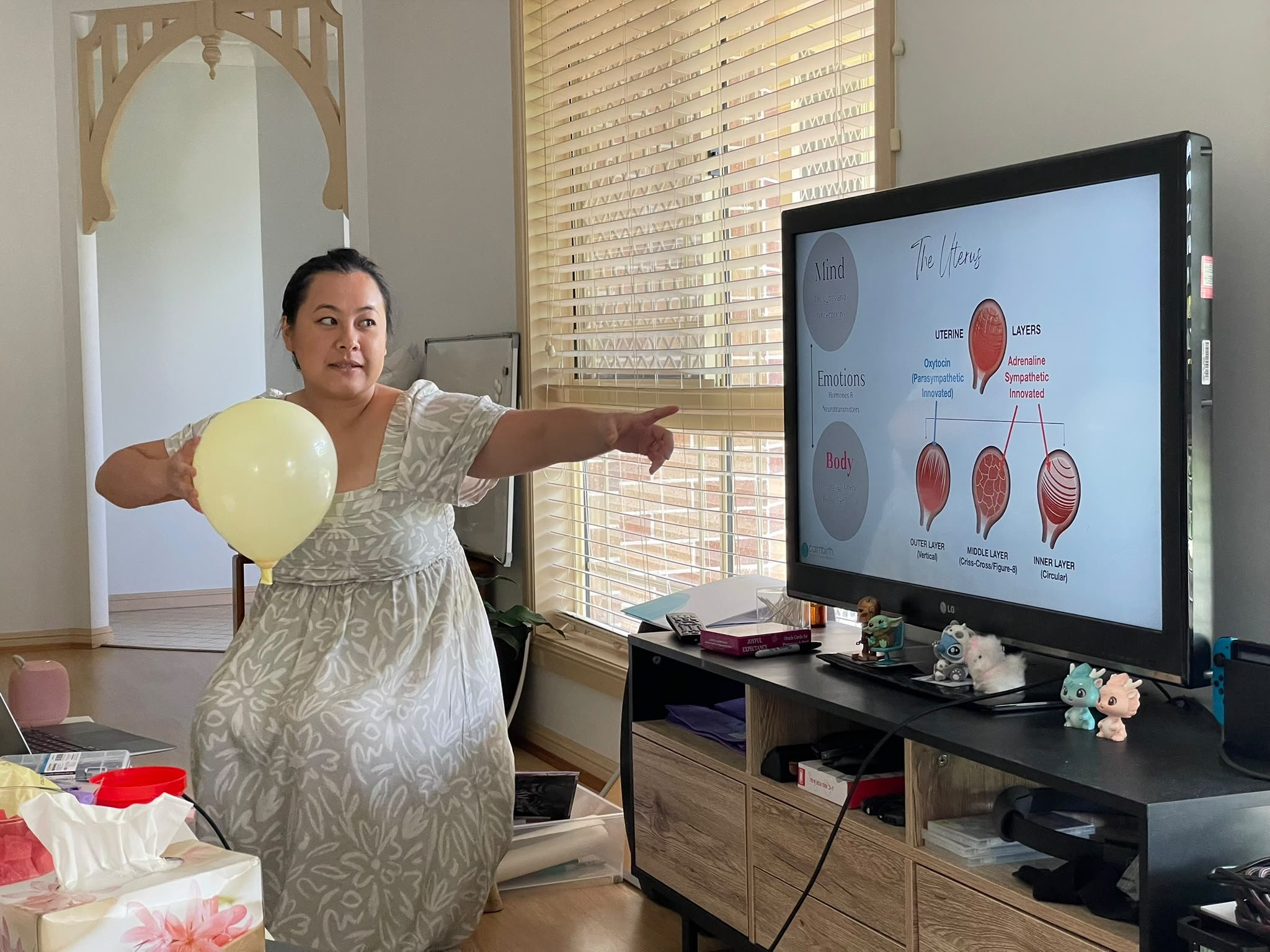 Woman in a patterned dress holding a yellow balloon and pointing at a screen displaying a diagram about the layers of the uterus. The room has a window with blinds, a black sideboard with plush animal toys, and various papers and objects on the table.