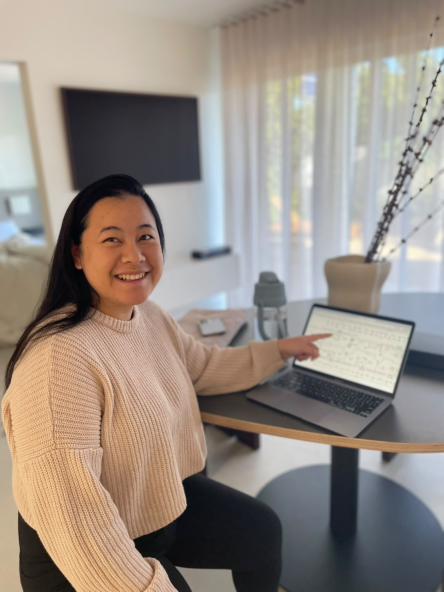 A smiling woman with long dark hair wearing a beige sweater sitting at a round table with a laptop, pointing at the screen, in a bright room with large windows and sheer curtains, a tall decorative vase with branches, and a TV mounted on the wall.