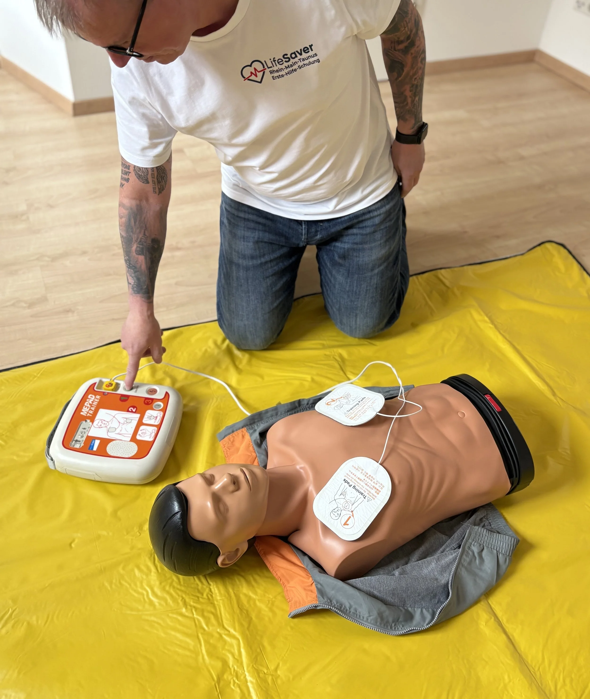 Ein Mensch übt an einem Dummy mit AED-Gerät in einem Rettungssimulator. Der Mensch trägt eine weiße Kappe und ein weißes T-Shirt mit einem Logo der Lebensretter. Der Dummy liegt auf einer gelben Unterlage auf dem Boden.