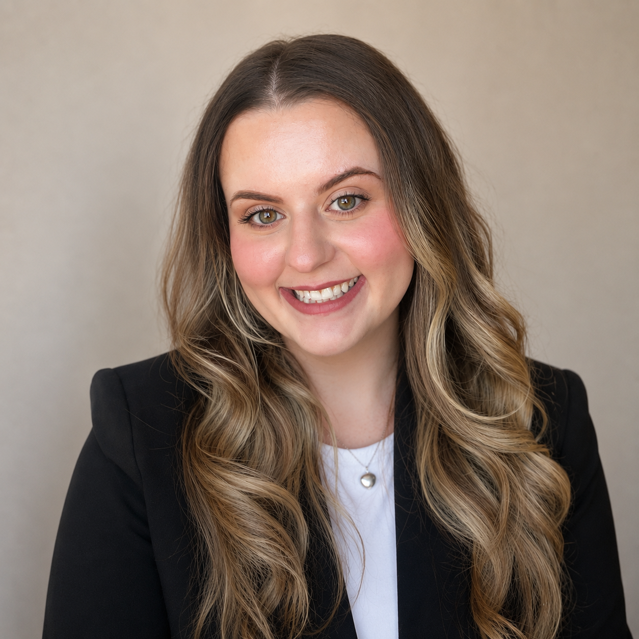 A young woman with long wavy blonde hair wearing a black blazer and a white top, smiling at the camera against a plain neutral background.
