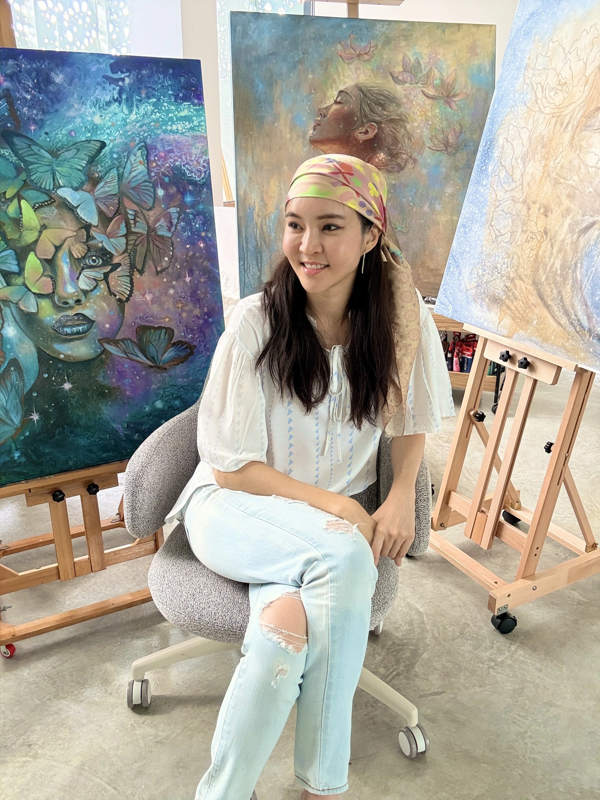 A woman with long dark hair wearing a beige and pink patterned headscarf, a white blouse, and ripped light blue jeans, sitting in an art gallery with paintings behind her. One painting depicts a woman's face surrounded by butterflies, and another shows a serene profile of a woman with flowers and butterflies around her.