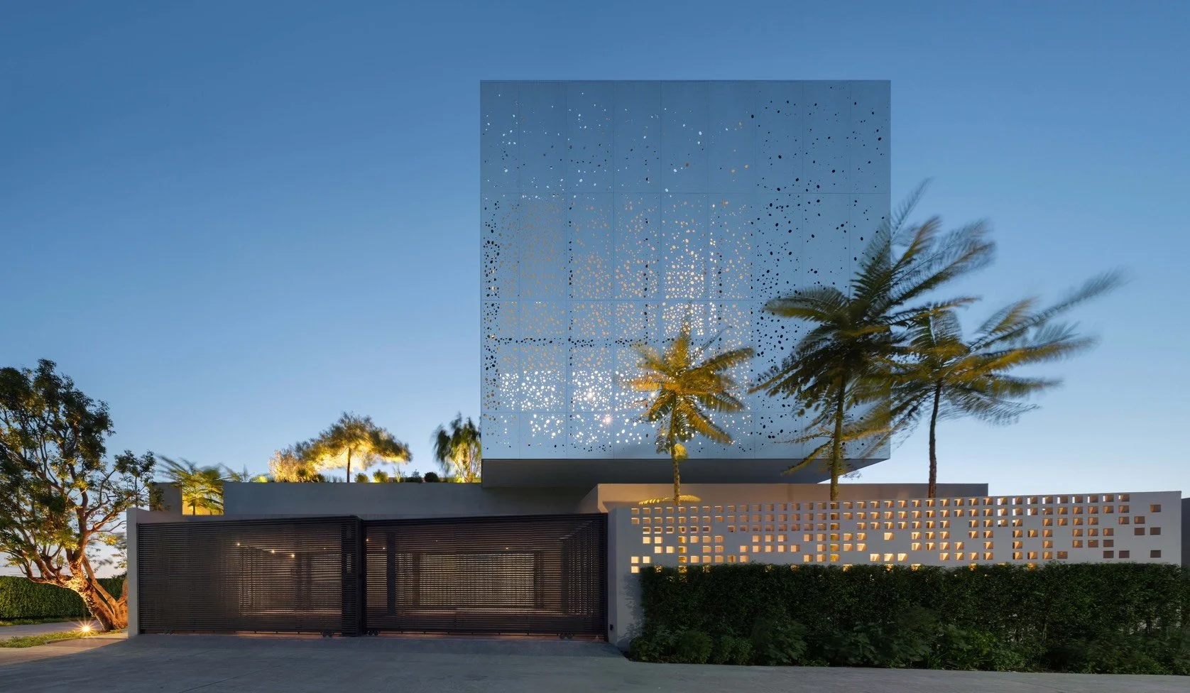 Modern building with perforated decorative facade, palm trees, a gated entrance, and a clear blue sky at dusk.
