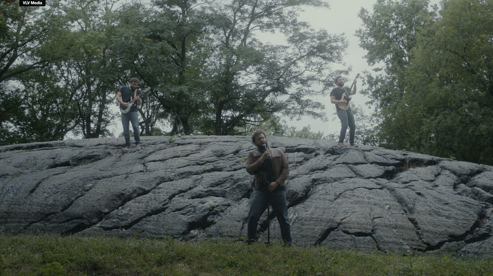 Three musicians perform outdoors on a large rocky surface, with trees in the background.