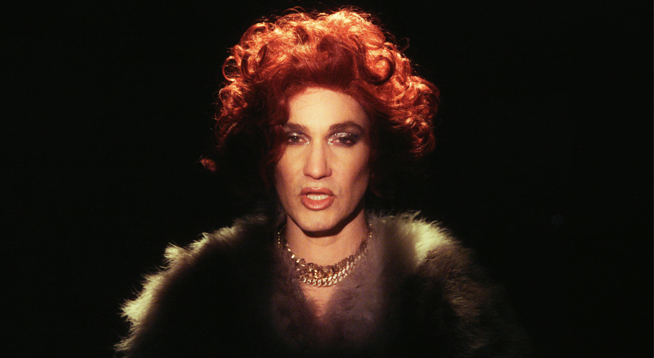 A woman with curly red hair, wearing dramatic makeup, gold jewelry, and a fur coat, appears to be singing or speaking, with a dark background.