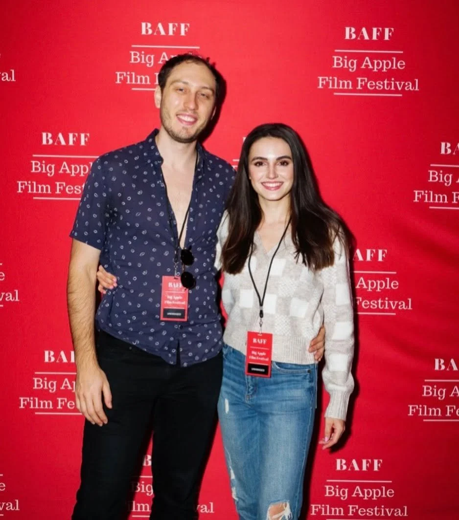 A young man and woman are posing together at the BAFF Big Apple Film Festival, standing in front of a red backdrop with white text promoting the event. The man is wearing a navy blue short-sleeve shirt with white polka dots, with sunglasses hanging around his neck. The woman is wearing a white sweater with a checkered pattern and distressed jeans. Both are smiling and have festival badges around their necks.
