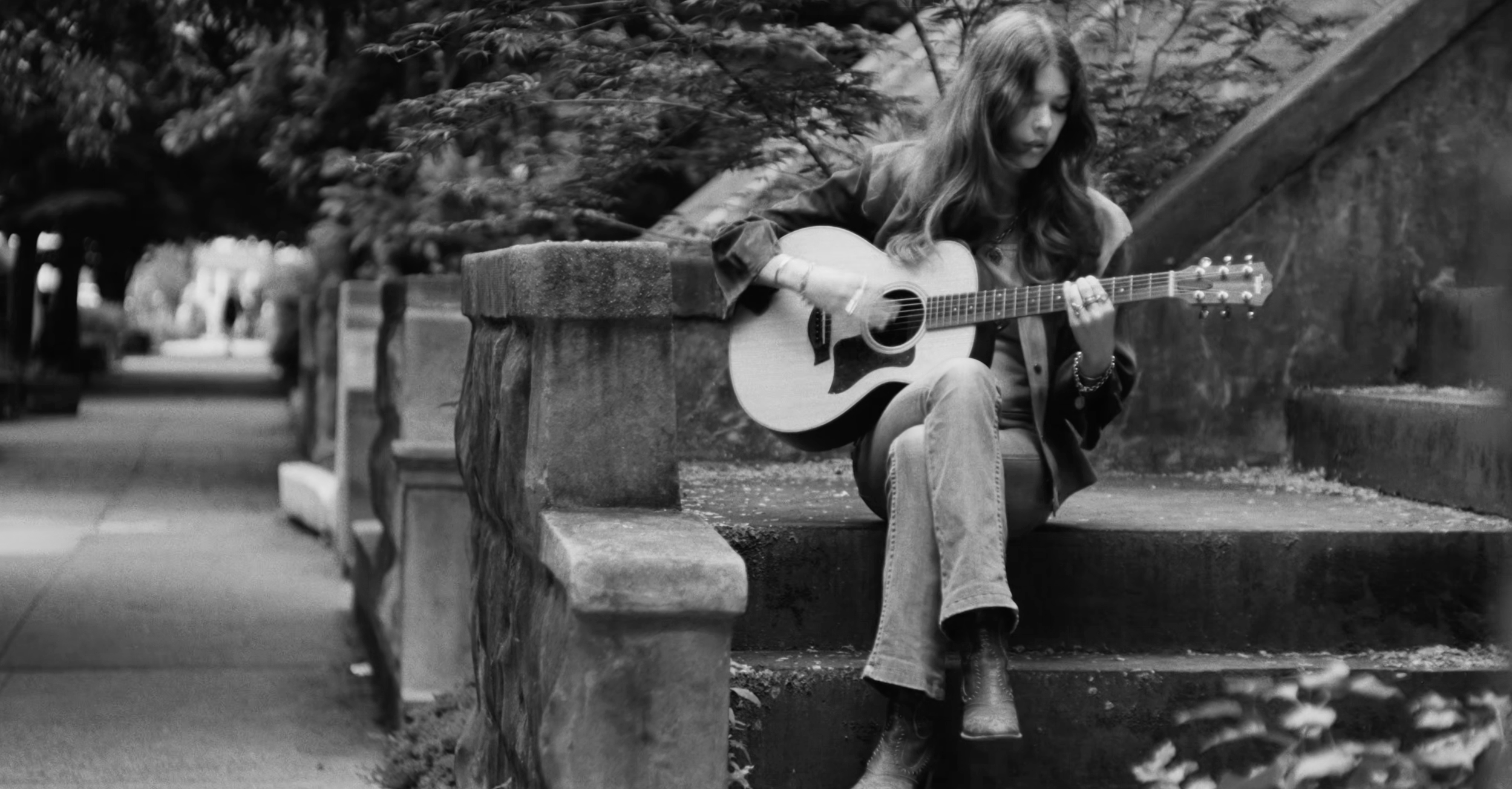 A young woman with long hair sitting on stone steps outdoors, playing an acoustic guitar.