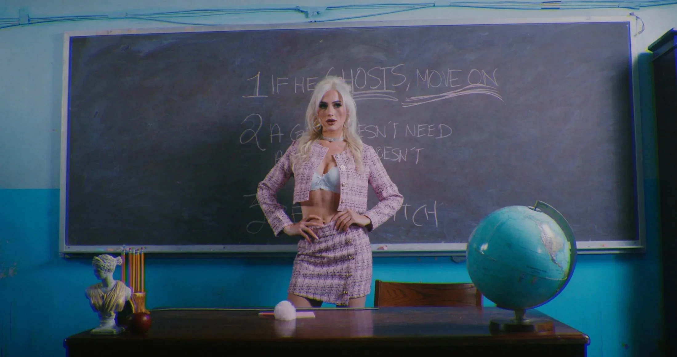 A woman with long blonde hair stands in front of a chalkboard in a classroom. The chalkboard has writing that says "If he ghost, move on" and "A guy isn't needed." There is a globe on a desk in the foreground, along with other classroom objects like a bust, a apple, and colored pencils.