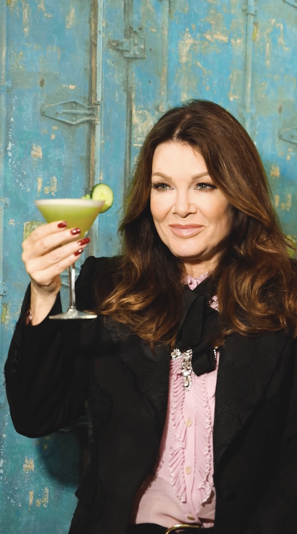 Woman with long brown hair holding a green cocktail with a cucumber slice, standing against a blue, distressed metal background.
