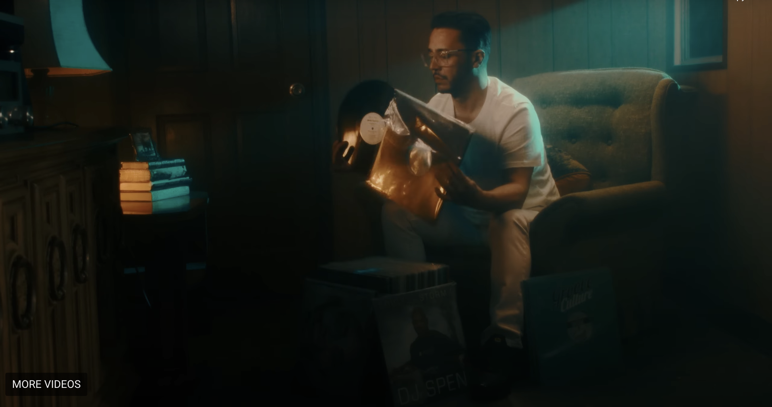 A man wearing glasses and a white shirt sitting on a sofa in a dimly lit room, examining a vinyl record album.
