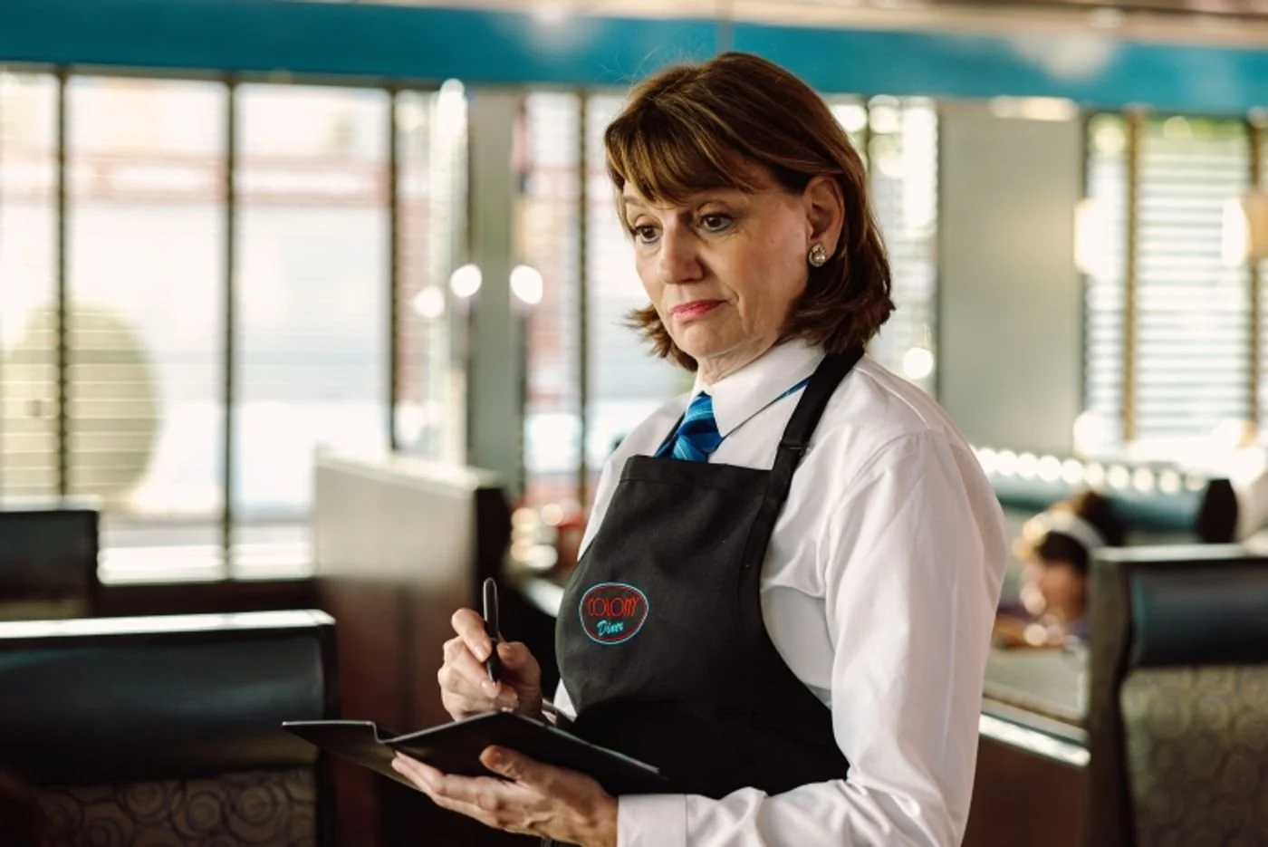 A middle-aged woman with brown hair, wearing a white shirt, blue tie, and black apron with a name tag, is inside a restaurant. She is holding a notepad and pen, appearing to take an order.