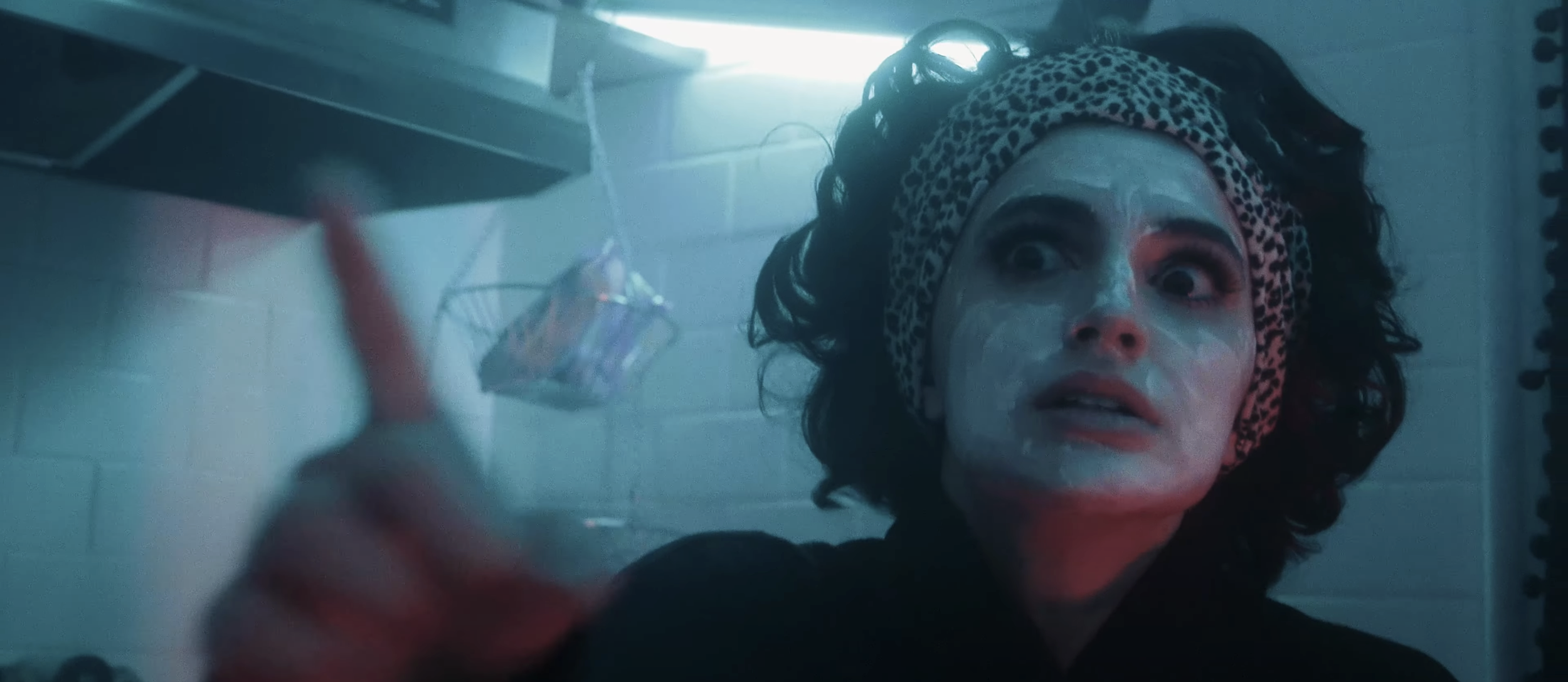Woman with purple face mask and leopard print headband looking up in a bathroom, reaching for a toiletries shelf.