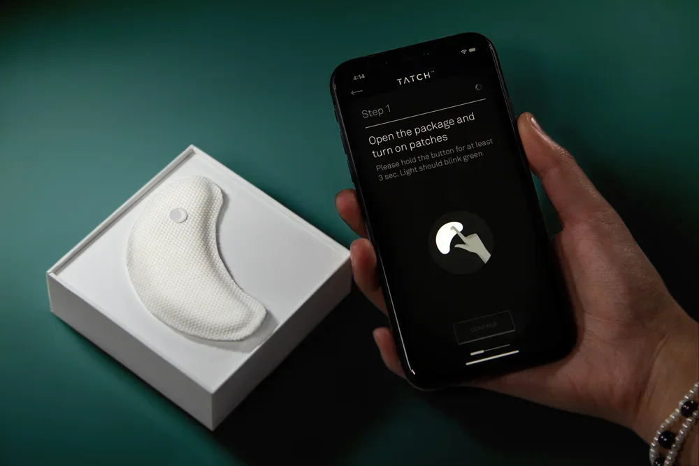 A person holding a smartphone displaying setup instructions for a TATCHI device, with a white, curved patch-like device in an open box on a green surface.