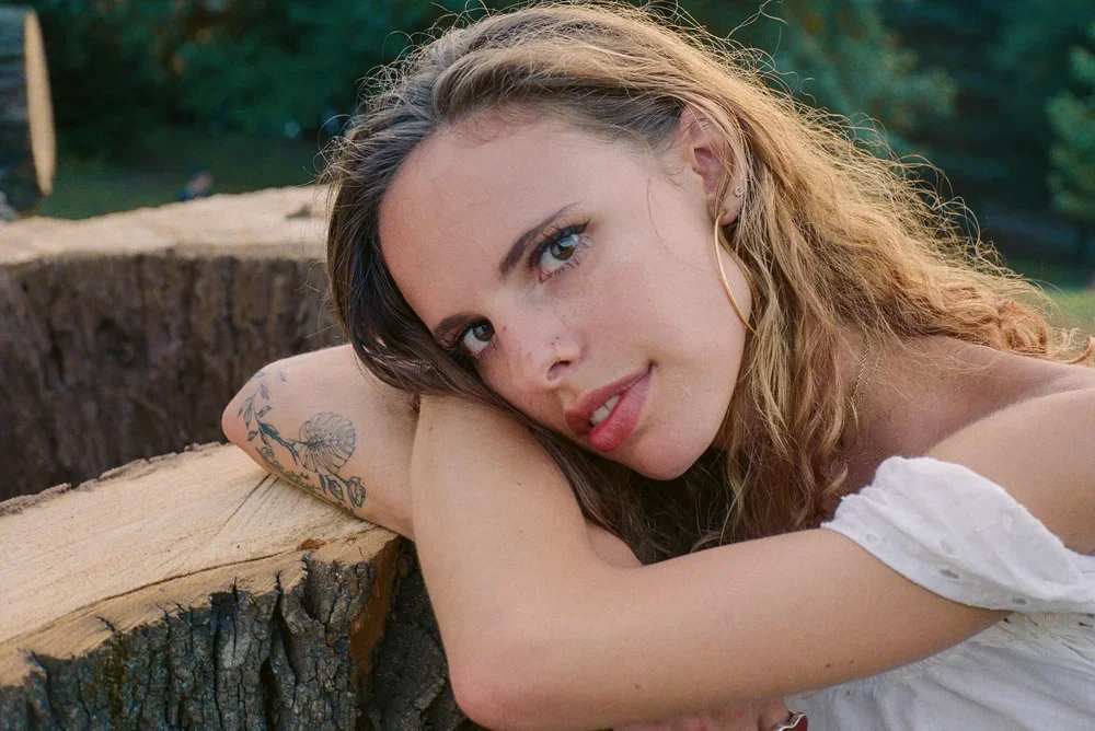 A young woman with long wavy hair and tattoos laying on a fallen tree trunk outdoors, looking at the camera with a slight smile.