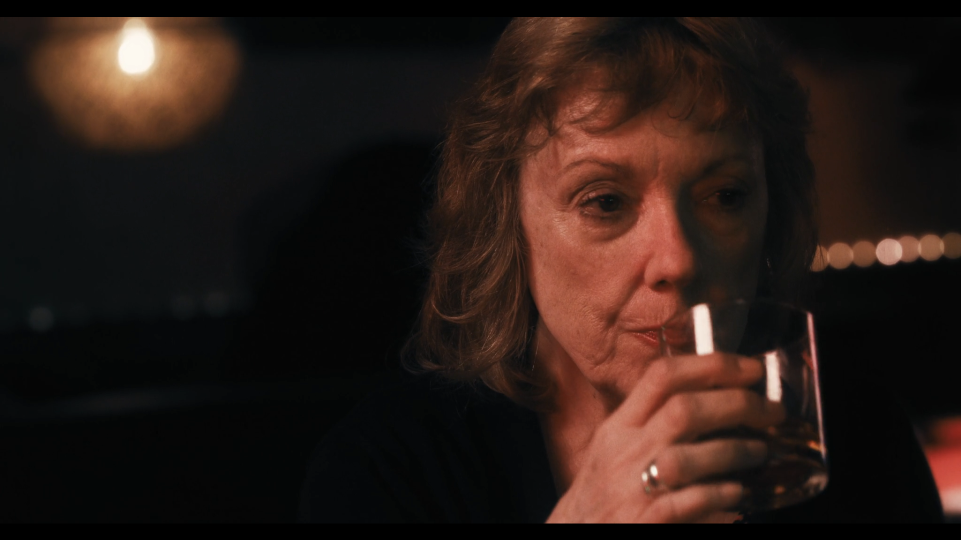 A middle-aged woman with short wavy brown hair drinking from a glass of dark-colored beverage in a dimly lit setting.