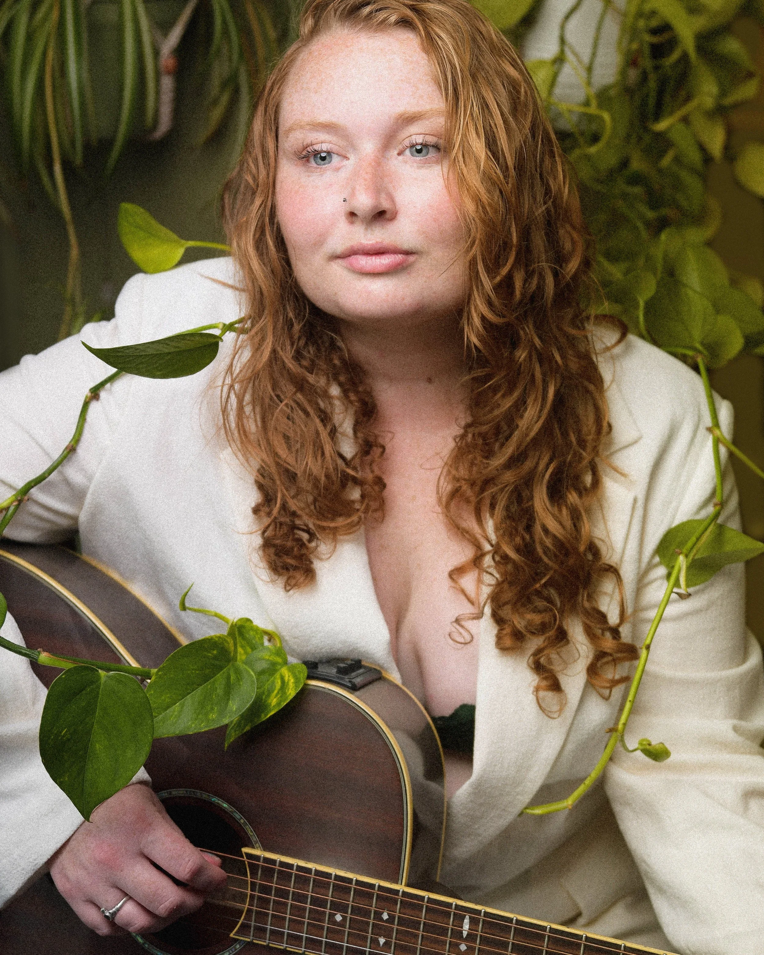 A woman with long, curly red hair and a nose piercing playing an acoustic guitar surrounded by green plants.