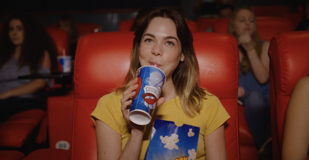 Young woman in a yellow shirt sitting in a movie theater, drinking soda from a large cup and holding popcorn.