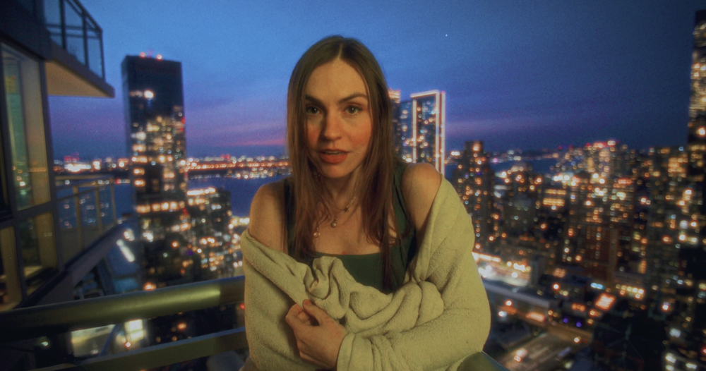 A woman with long red hair standing on a balcony overlooking a city at dusk with illuminated buildings and a river.