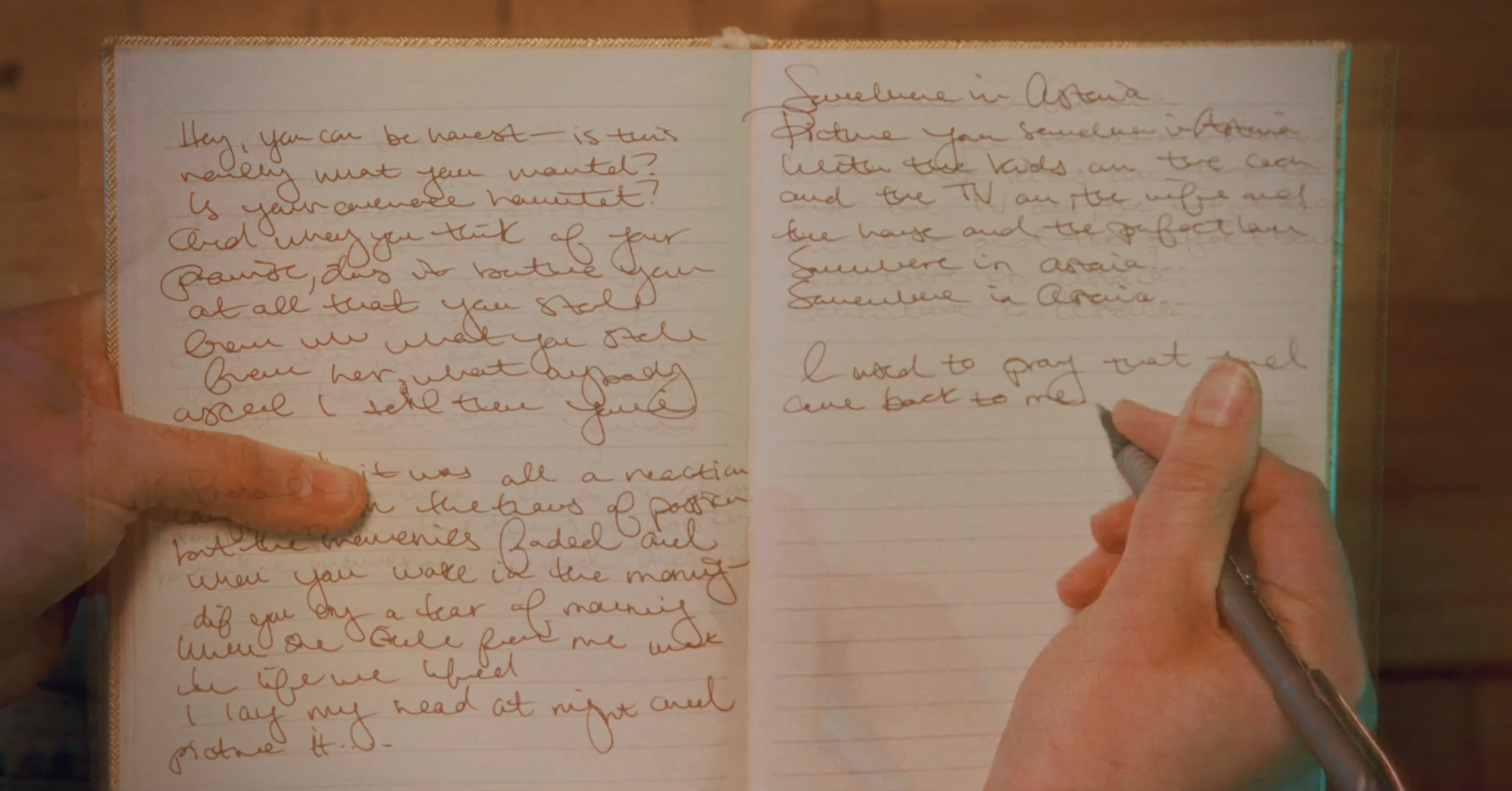 Close-up of a handwritten journal or notebook with cursive writing, held in someone's hands, with a person writing on the right page.