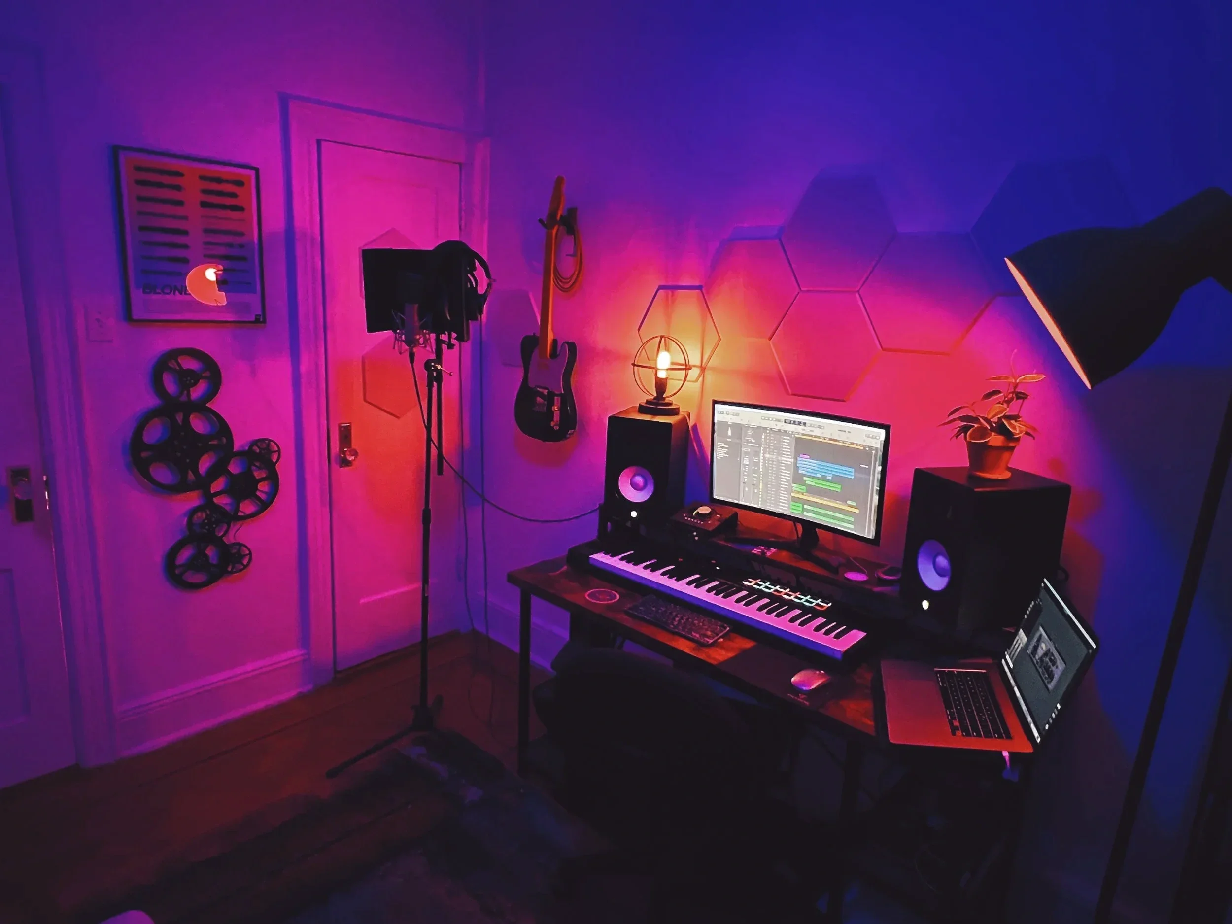 A music studio corner with purple, pink, and blue lighting. Contains a computer monitor, a keyboard, a MIDI controller, a speaker, a potted plant, a music stand, and a guitar hanging on the wall. There are film reels mounted on the wall.