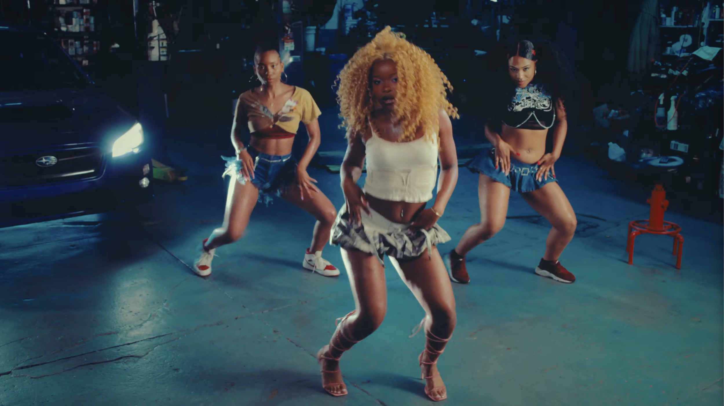 Three women dance in a dimly lit garage or workshop, with one woman with curly blonde hair in a cream top and patterned shorts in the center, and two women with long dark hair wearing crop tops and shorts on either side.