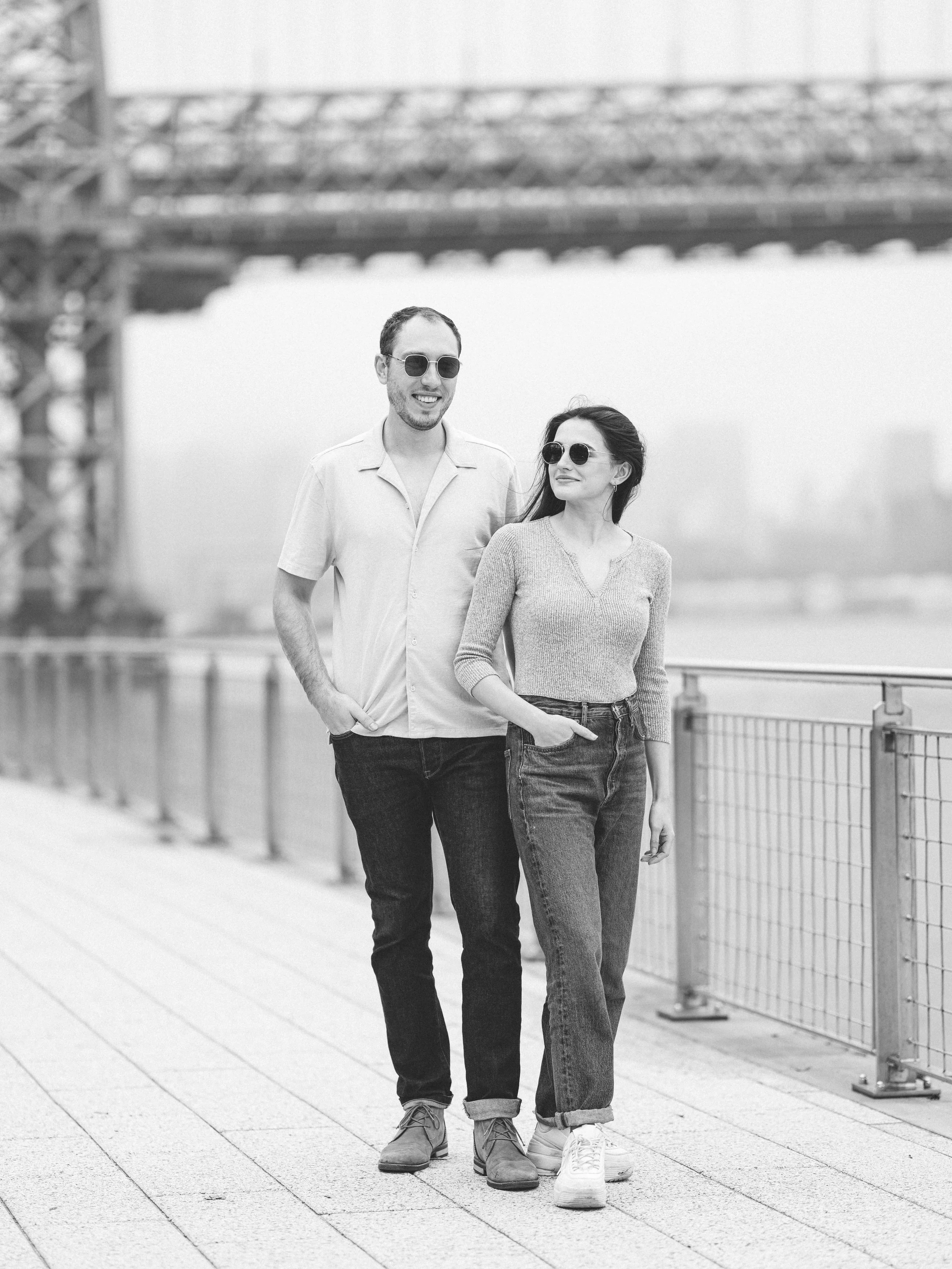 A black and white photo of a man and woman walking together outdoors on a paved area near a railing. They are both wearing sunglasses and casual clothing, with the man smiling and the woman looking slightly to the side.