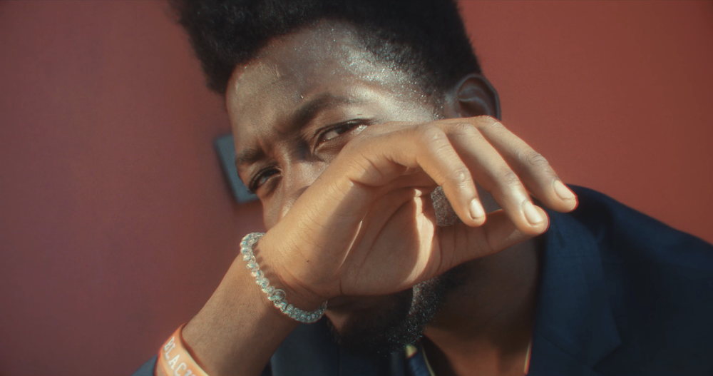 Close-up of a man with dark curly hair and a beard, wearing a blue shirt, covering part of his face with his hand, wearing a bracelet and wristband, standing against a pinkish-red wall.