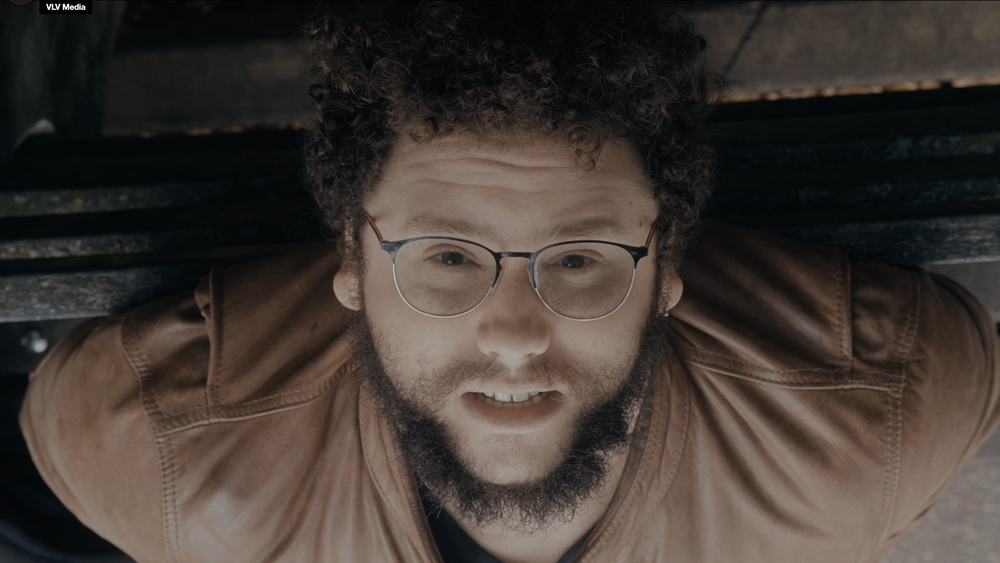 A man with curly hair, glasses, and a beard looking up at the camera, with a slight smile, wearing a brown jacket.