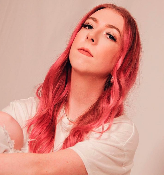 A young woman with long, wavy pink hair, wearing a white t-shirt, looking confidently at the camera against a plain background.