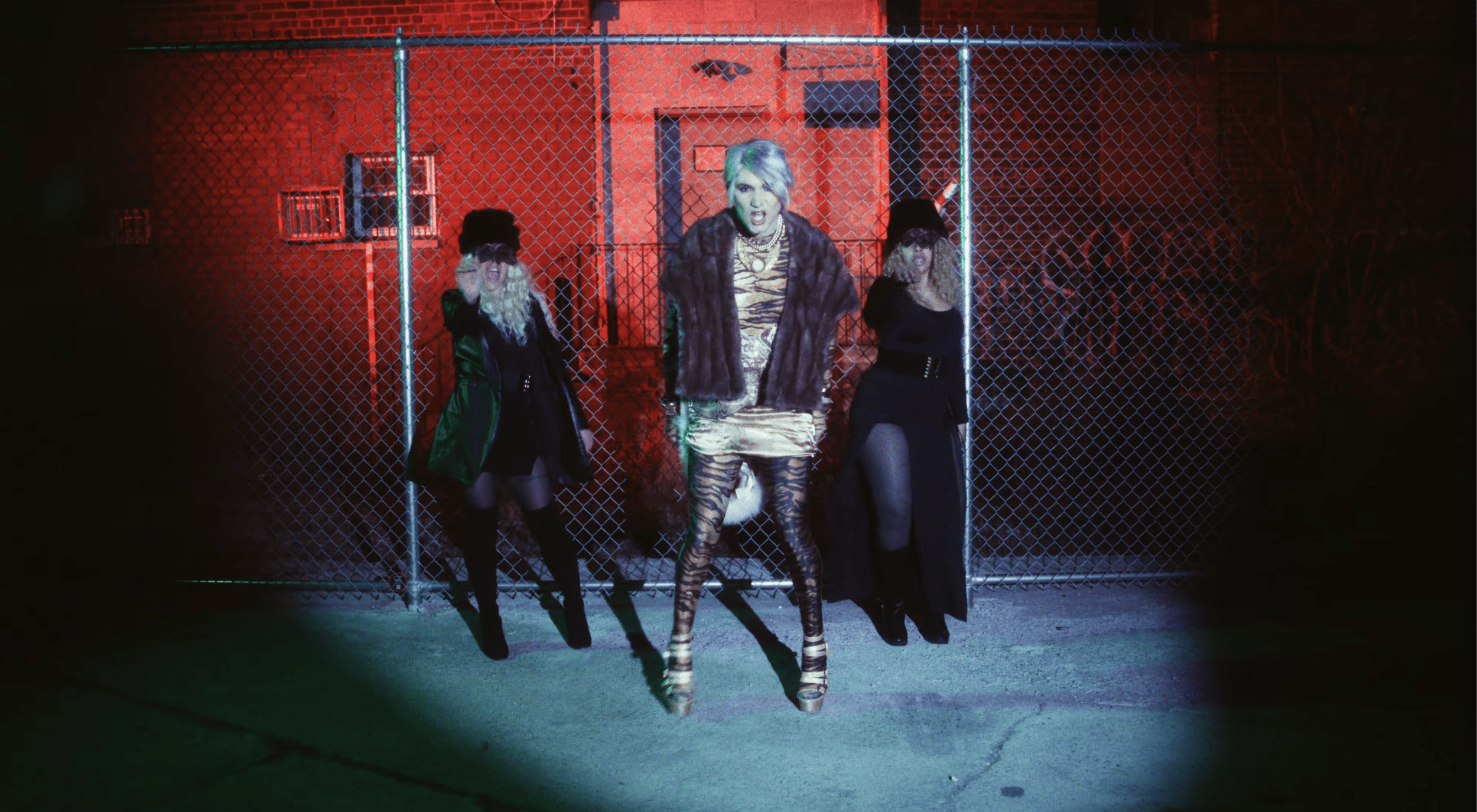 A woman with short, platinum blonde hair and animal print tights stands in front of a chain-link fence at night, flanked by two women with long, curly hair wearing black outfits and hats, illuminated by colorful neon lights, with a red building in the background.