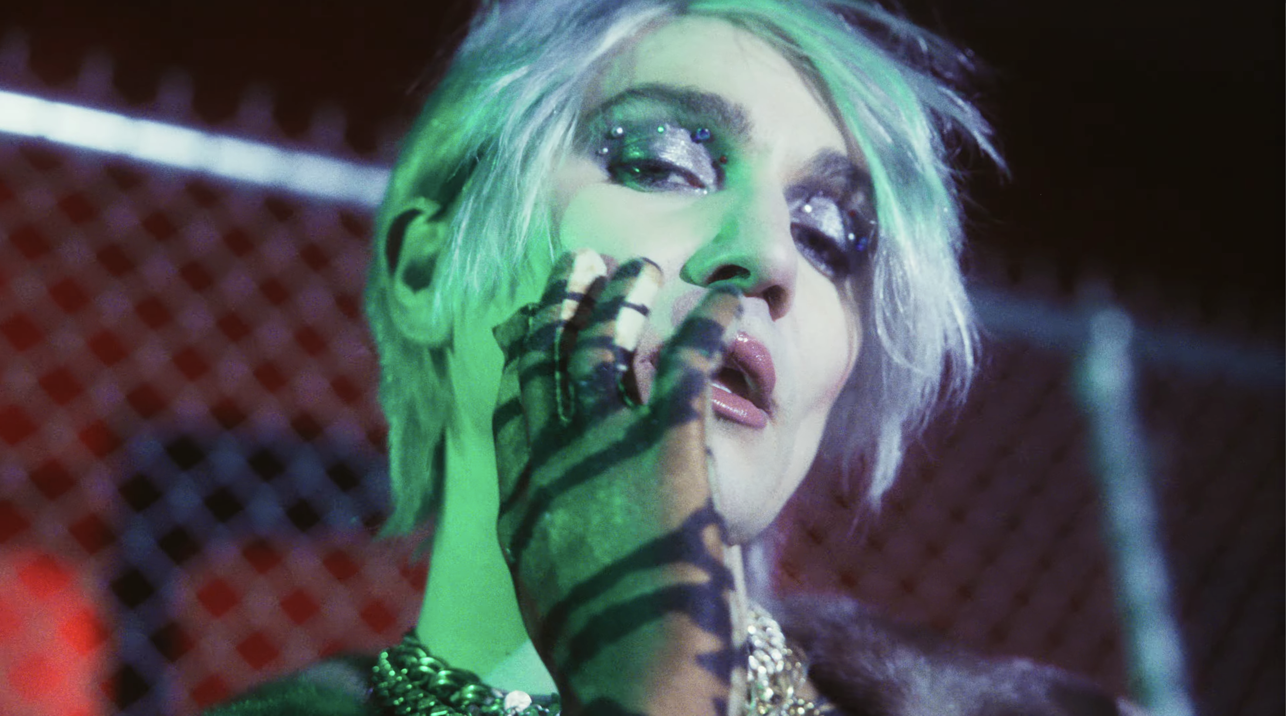 A woman with platinum blonde hair styled in a messy look, wearing dark makeup and multiple earrings, touching her face with gloved hands in a dim, colorful, and possibly neon-lit environment.