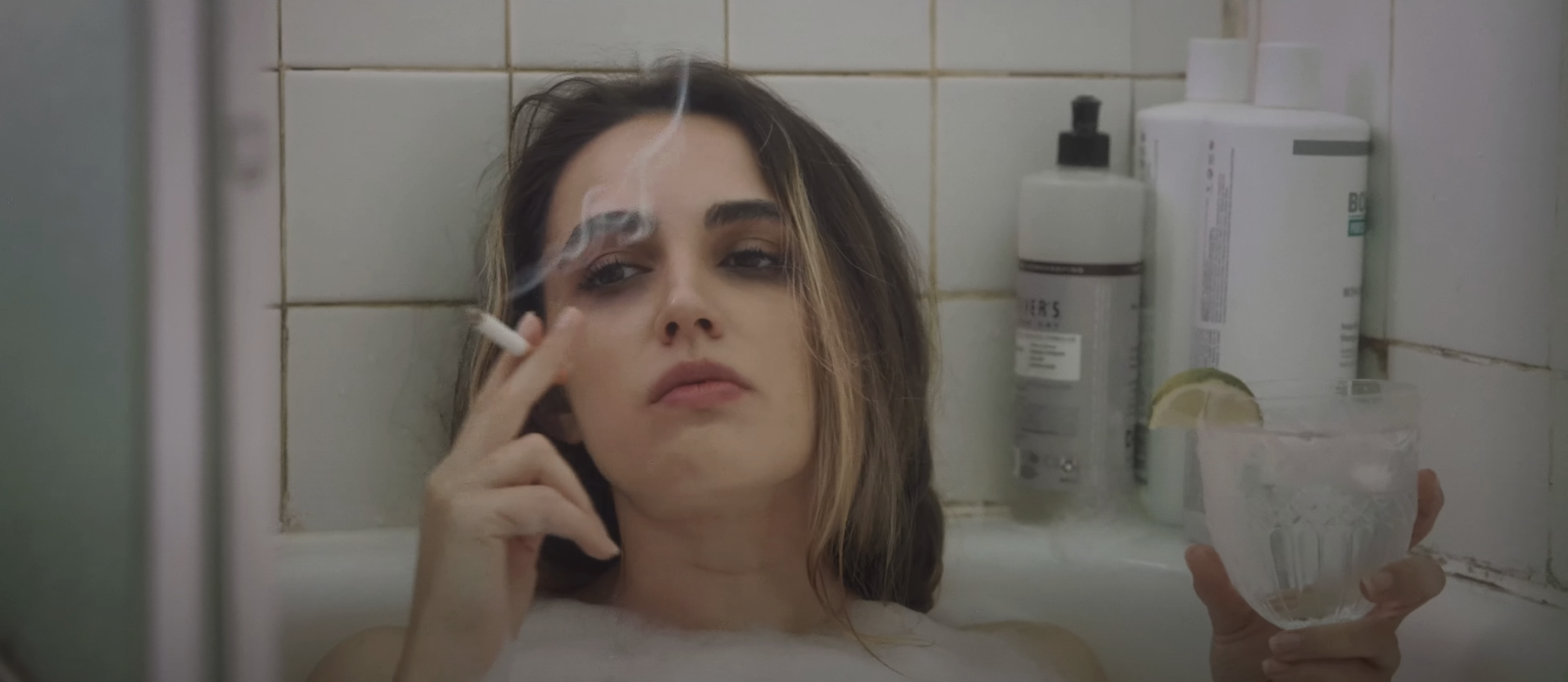 A woman with brown hair and makeup, smoking a cigarette, relaxing in a bathtub filled with water, holding a glass of ice water with lemon slices.