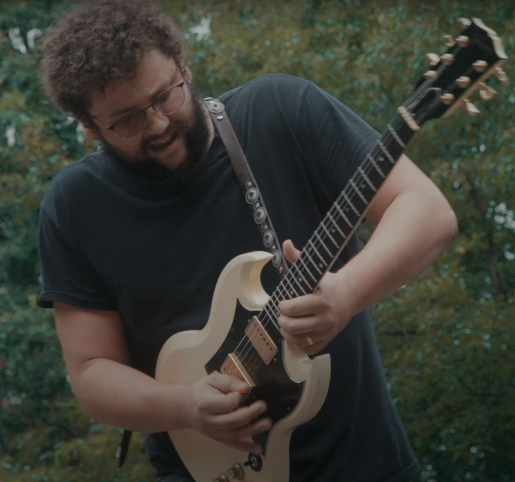 A man with curly hair, glasses, and a beard playing an electric guitar outdoors, with trees in the background.