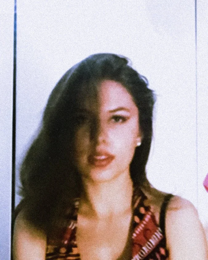A woman with dark hair styled voluminously, wearing red lipstick and a patterned dress, standing against a light-colored background.