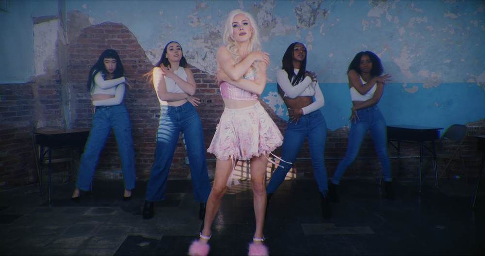 A woman with blonde hair in a pink outfit standing in front of four women with different hairstyles and skin tones, dressed in white tops and blue jeans, in a room with a partly exposed brick wall and distressed paint.