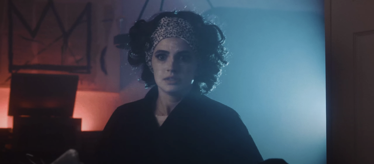A woman with curly hair, wearing a leopard print headband and dark clothing, sits in a dimly lit room with blue and red lighting, looking directly at the camera.
