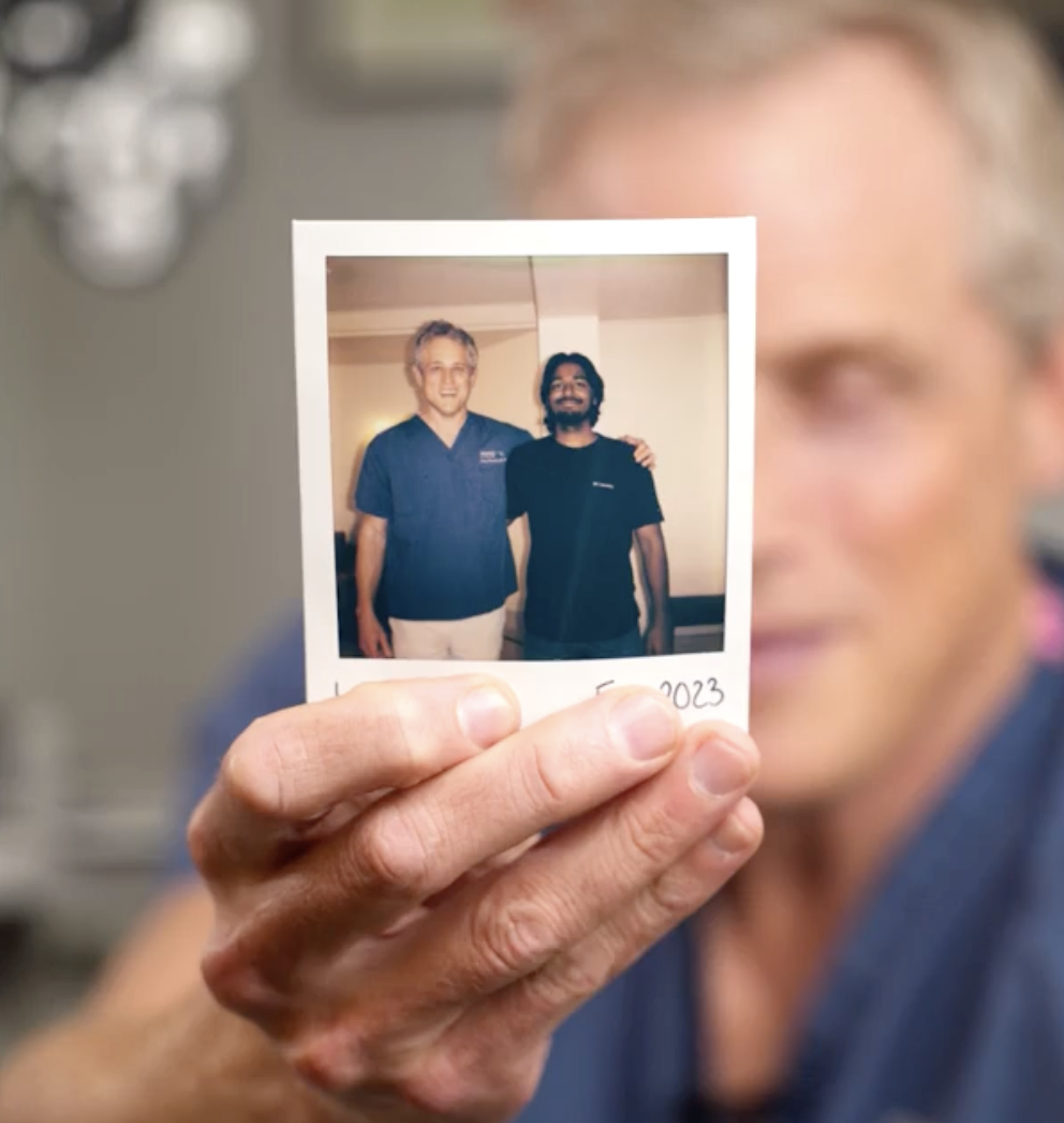 A woman holding a small printed photograph of two men, one in medical scrubs, smiling and standing together in an indoor setting.