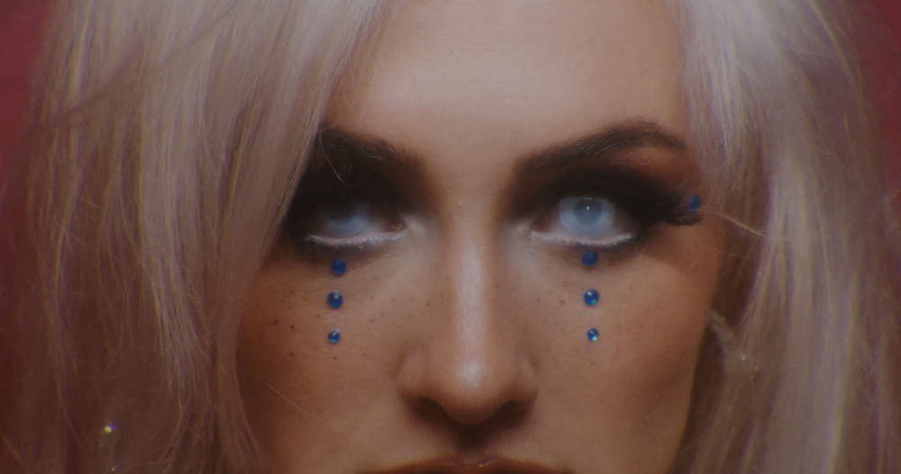 Close-up of a woman with light purple hair and dramatic eye makeup with blue rhinestones underneath her eyes, looking slightly upward.