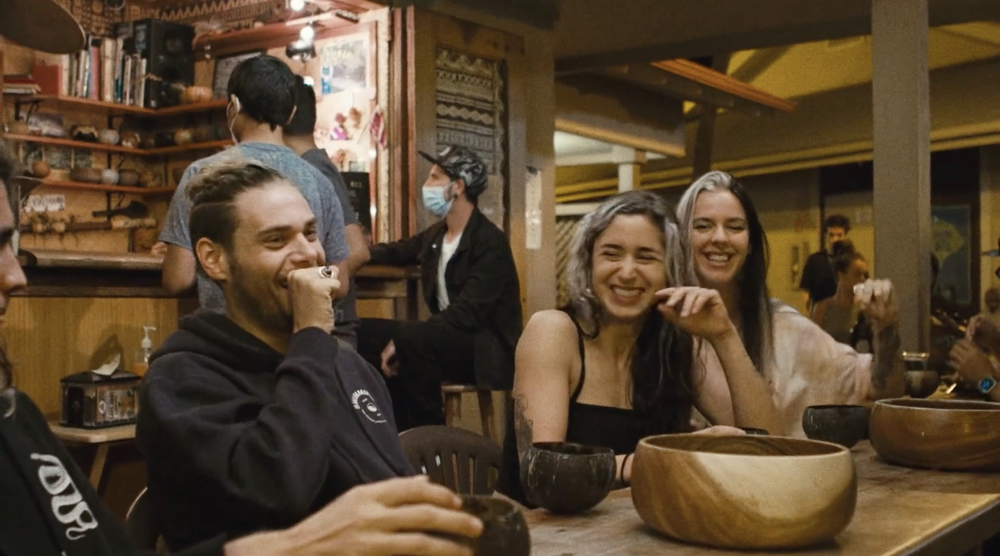 Group of young adults sitting at a wooden table in a cozy, rustic-style restaurant, smiling and laughing, with bowls on the table. In the background, a person wearing a mask stands near a bar area.