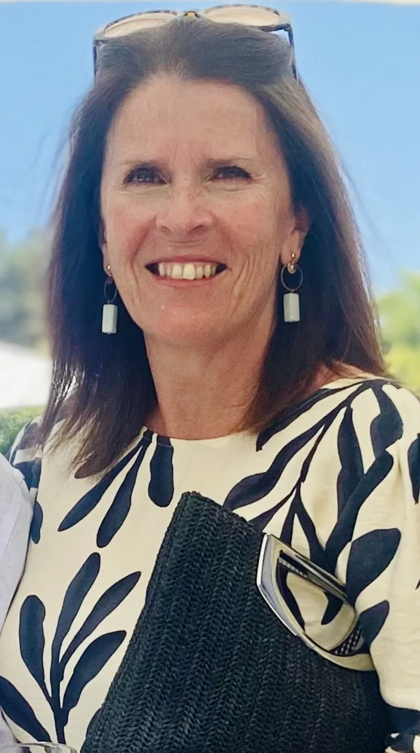 A smiling woman with dark hair, wearing sunglasses on her head, a black and white patterned top, earrings, and holding a black woven bag outdoors with blue sky and trees in the background.