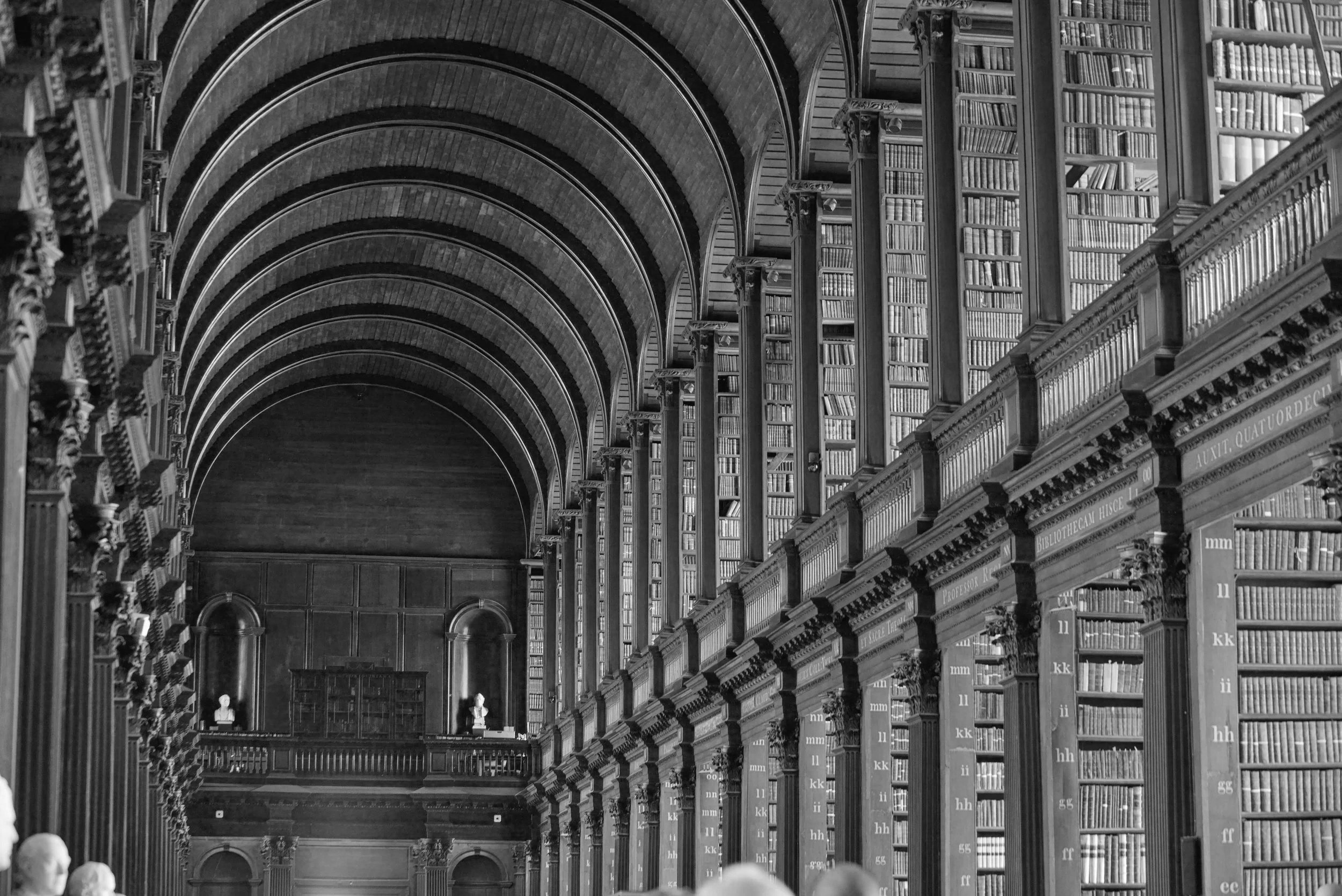Black and white photo of a grand historic library with high arched ceilings, wooden bookshelves filled with books, and bust sculptures along the lower shelves.