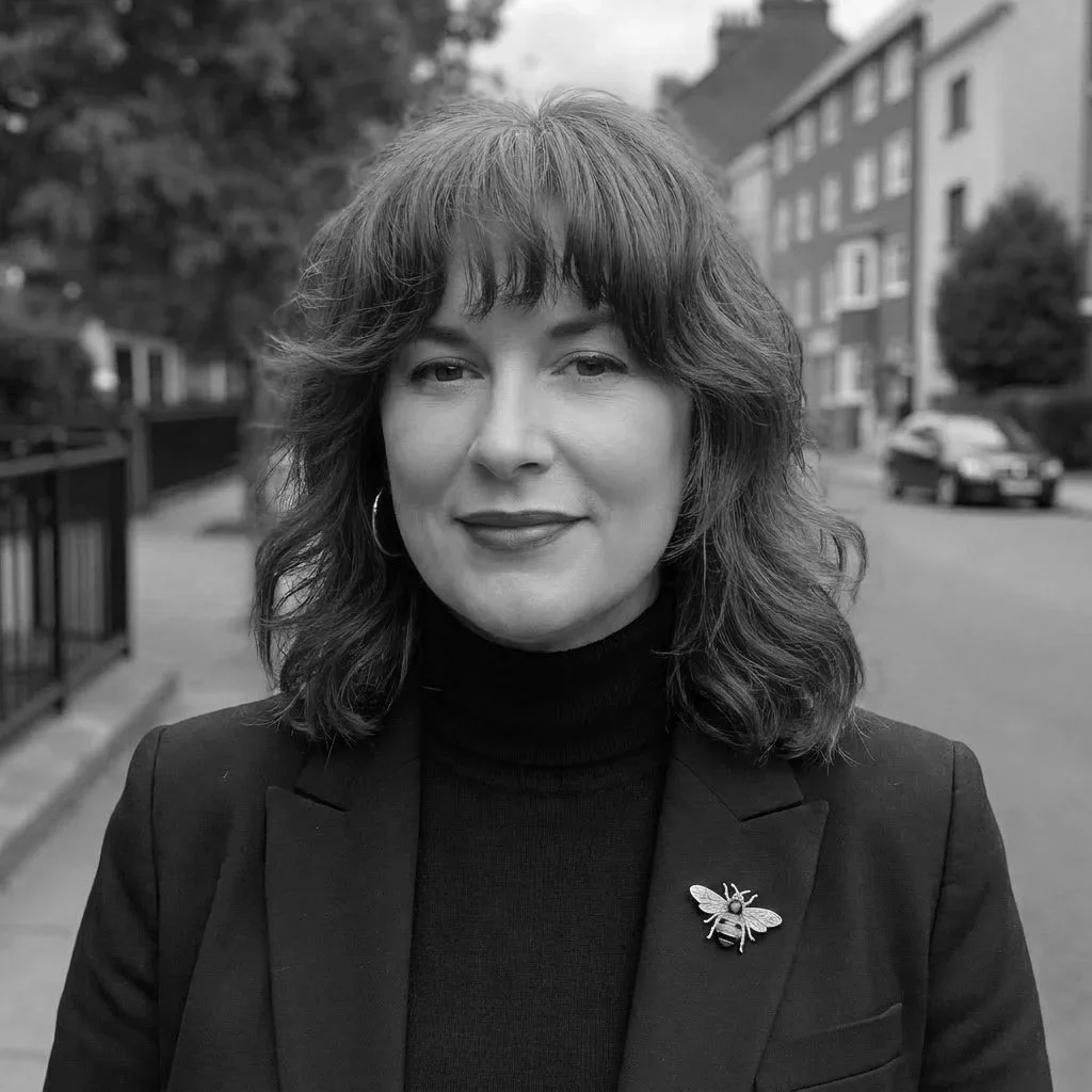 Black and white photograph of a woman with shoulder-length wavy hair, wearing a dark blazer with a bee brooch, standing outdoors on a street with buildings, trees, and parked cars in the background.