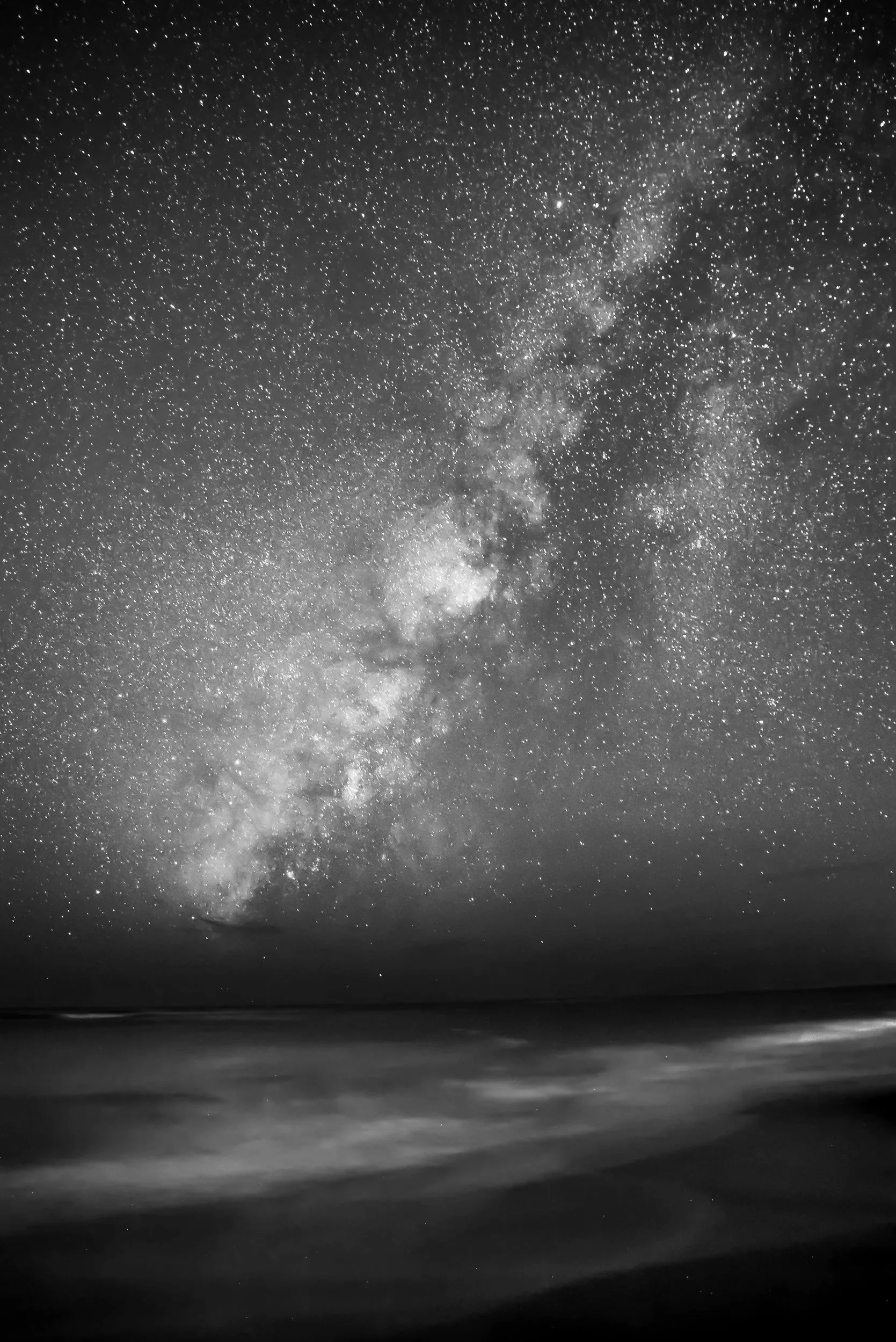 Black and white photo of the night sky filled with stars and the Milky Way galaxy visible, above a calm body of water.