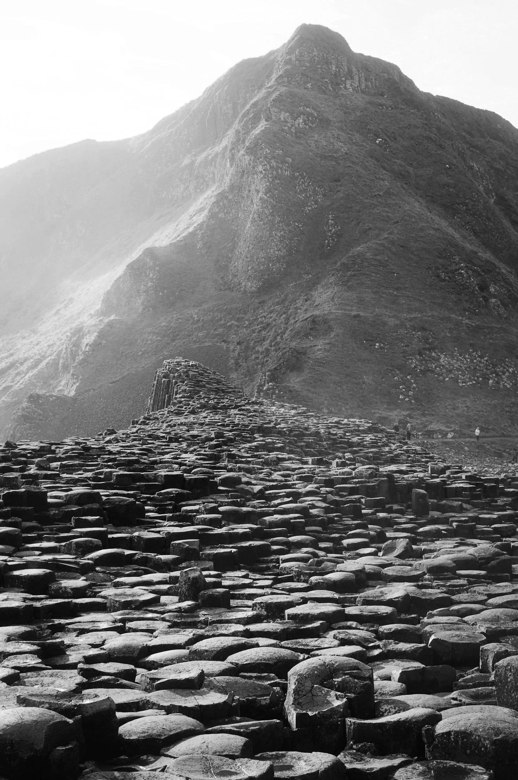 A black and white photo of Giants Causeway, a natural rock formation with hexagonal basalt columns at the foot of a mountain.