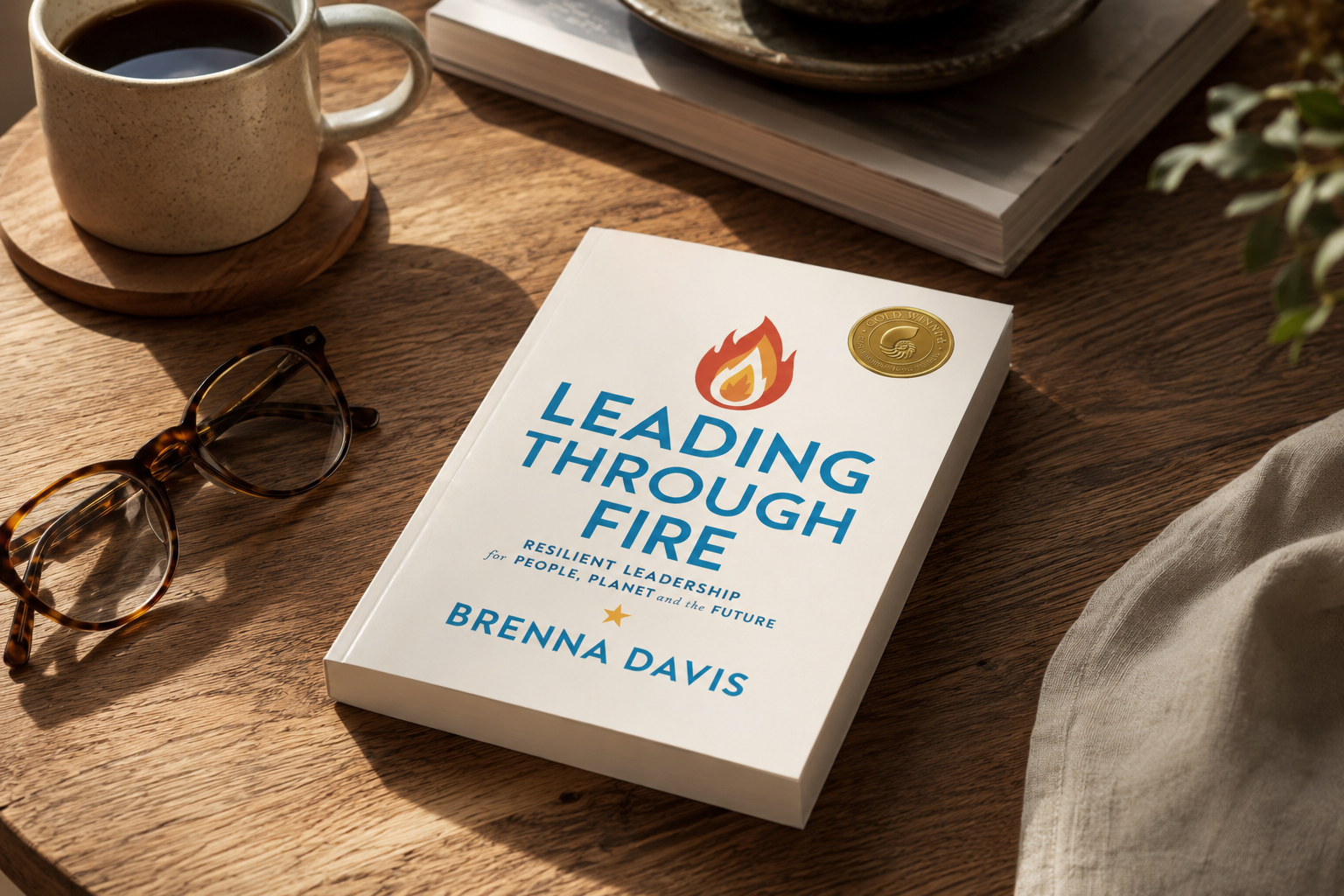 Book titled 'Leading Through Fire' by Brenna Davis on a wooden table with sunglasses, a cup of coffee, a plant, and a stack of magazines.