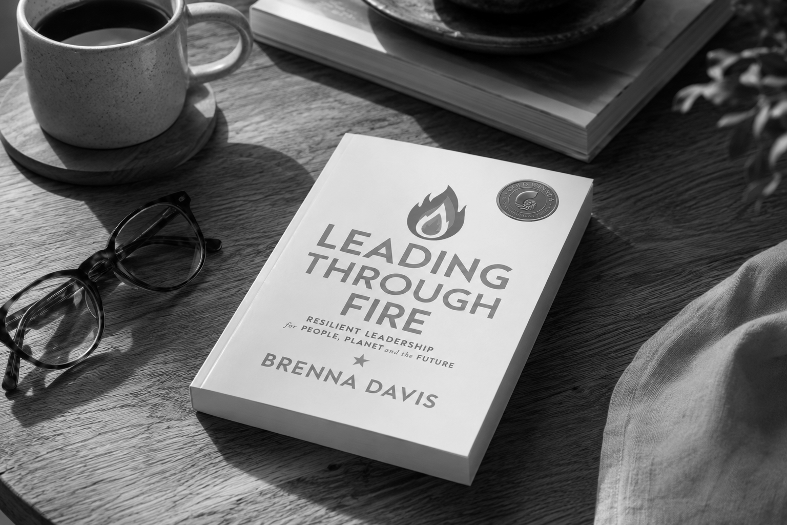 Black and white photo of a book titled 'Leading Through Fire' by Brenna Davis on a wooden table. Nearby are a mug of coffee on a small round tray, a pair of glasses, an open magazine, and part of a person's arm and cloth.