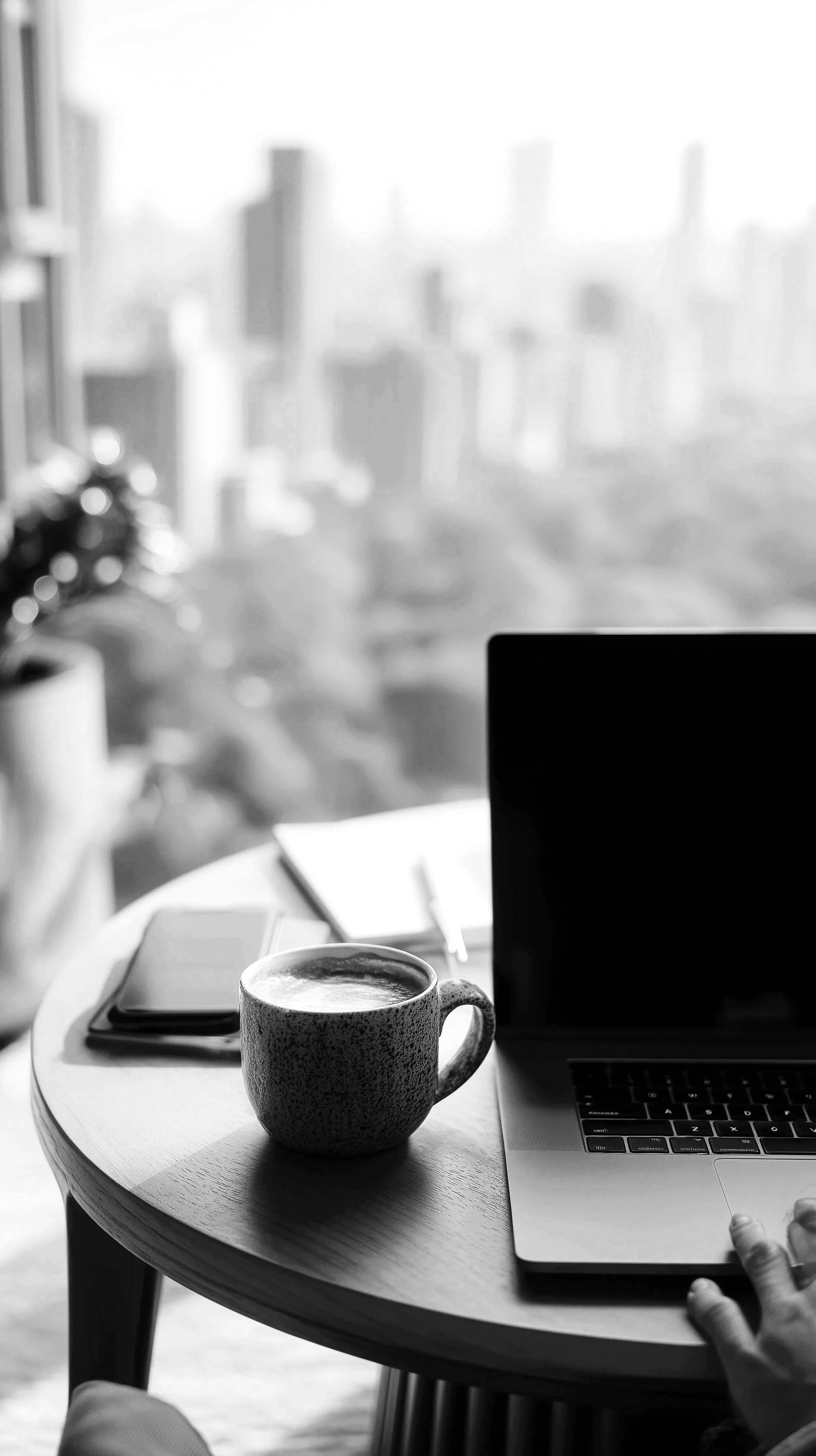 Black and white photograph of a coffee mug, a laptop, a notebook, and a pen on a round wooden table in a bright room with large windows and city skyscrapers in the background.